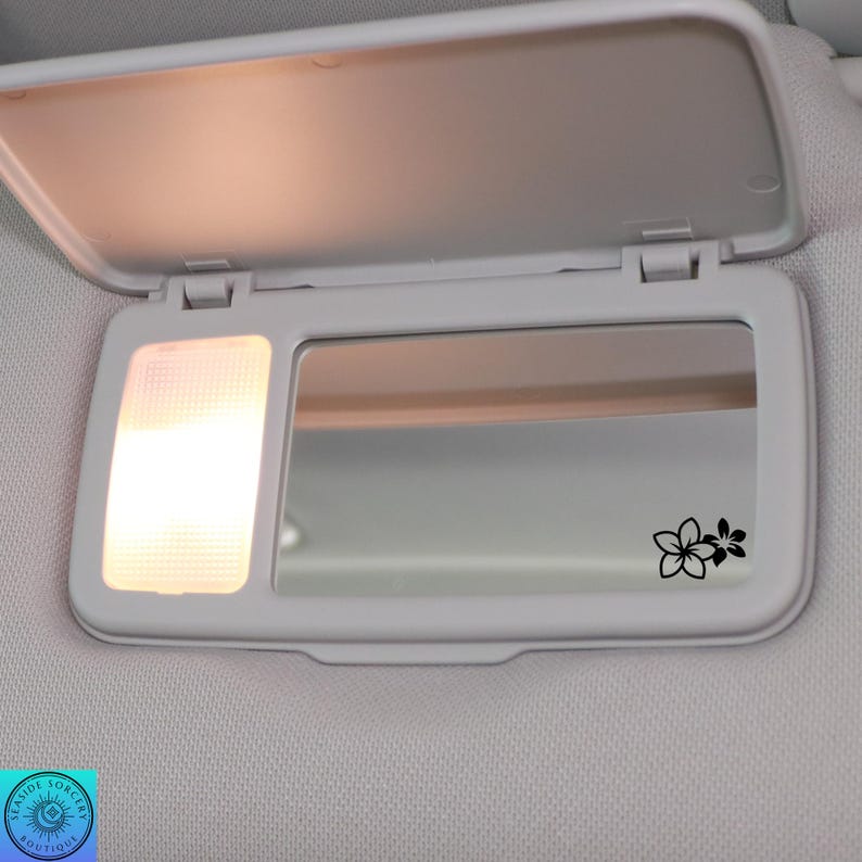 Pu&ograve; includere: Una visiera parasole bianca per auto con specchietto e piccola luce. Lo specchietto presenta un motivo floreale nero. La visiera &egrave; aperta e la luce &egrave; accesa, illuminando l'interno.