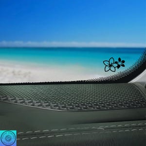 Pu&ograve; includere: Un adesivo floreale nero &egrave; applicato al finestrino di un'auto, con una scena di spiaggia e oceano sullo sfondo. L'adesivo presenta due fiori stilizzati. Il cruscotto dell'auto &egrave; visibile in primo piano, con una superficie strutturata e cuciture bianche.
