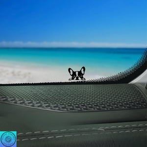 May include: A black and white French Bulldog decal peeking over a dark dashboard. The background features a bright blue sky, ocean, and sandy beach. The decal is a cute, decorative accessory.