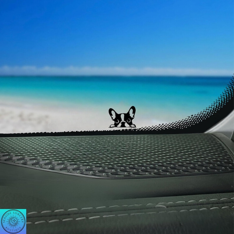May include: A black and white French Bulldog decal peeking over a dark dashboard. The background features a bright blue sky, ocean, and sandy beach. The decal is a cute, decorative accessory.
