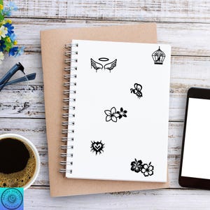 Pu&ograve; includere: Un quaderno a spirale bianco con varie illustrazioni nere, tra cui ali d'angelo, una gabbia per uccelli, un'ape, fiori, un cuore e ibisco. Il quaderno &egrave; su un blocco note marrone, con caff&egrave;, occhiali e un telefono.