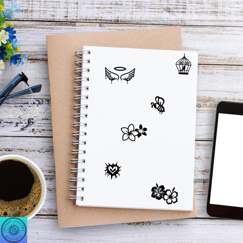 Pu&ograve; includere: Un quaderno a spirale bianco con varie illustrazioni nere, tra cui ali d'angelo, una gabbia per uccelli, un'ape, fiori, un cuore e ibisco. Il quaderno &egrave; su un blocco note marrone, con caff&egrave;, occhiali e un telefono.