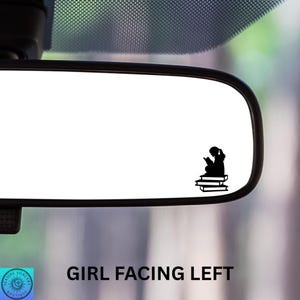 May include: Black vinyl decal of a girl reading, seated on a stack of books. The text "GIRL FACING LEFT" is below the rearview mirror. The decal is designed for application on a car window or similar surface.
