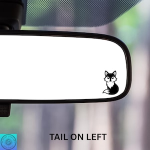 May include: A black and white fox decal is affixed to a car's rearview mirror. The decal features a stylized fox with its tail on the left. The text "TAIL ON LEFT" is below the mirror.