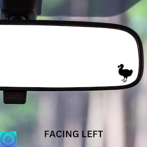 May include: A black silhouette of a dodo bird facing left, placed on a rearview mirror in a car. The mirror is reflecting a blurry green background.