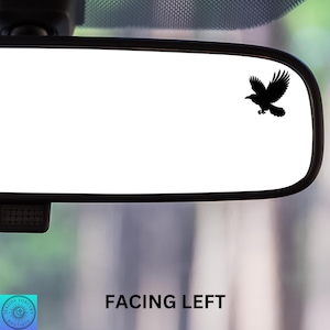 May include: A black silhouette of a bird facing left, placed on a white rearview mirror in a car.