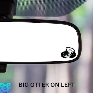 May include: A black and white vinyl decal of two otters cuddling, placed on a car's rearview mirror. The text "BIG OTTER ON LEFT" is at the bottom. The decal is a simple, graphic design.