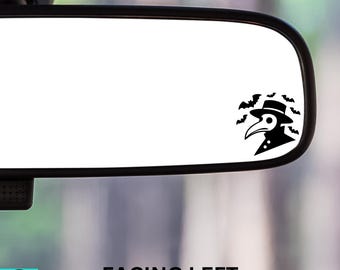 May include: A black and white car rearview mirror with a sticker of a plague doctor with a beak mask and hat, surrounded by bats. The text "FACING LEFT" is printed below the mirror.
