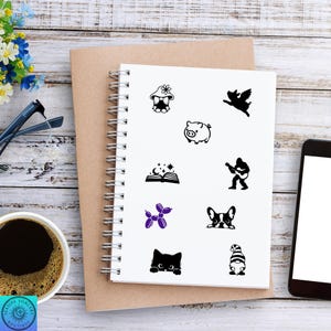 May include: A spiral-bound notebook with various black and purple graphic stickers. The stickers include a gnome, pig, angel, open book, guitar player, dog, cat, and balloon animal. A cup of coffee, glasses, and a phone are also visible.