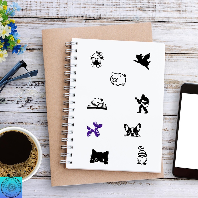 May include: A spiral-bound notebook with various black and purple graphic stickers. The stickers include a gnome, pig, angel, open book, guitar player, dog, cat, and balloon animal. A cup of coffee, glasses, and a phone are also visible.