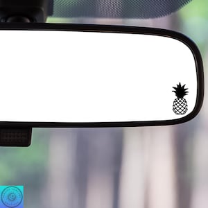 May include: A black pineapple decal is affixed to the lower right corner of a car's rearview mirror. The mirror is rectangular with rounded corners and a black frame. The background is blurred, suggesting an outdoor setting.