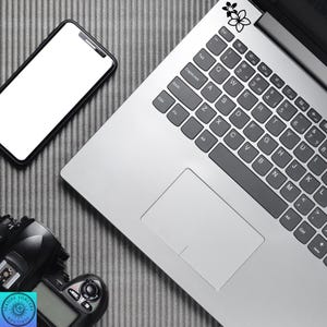 Pu&ograve; includere: Un laptop con un adesivo floreale in bianco e nero, uno smartphone e una fotocamera digitale sono disposti su una superficie grigia strutturata. La tastiera del laptop &egrave; visibile e la fotocamera ha un logo Seaside Sorcery Boutique.