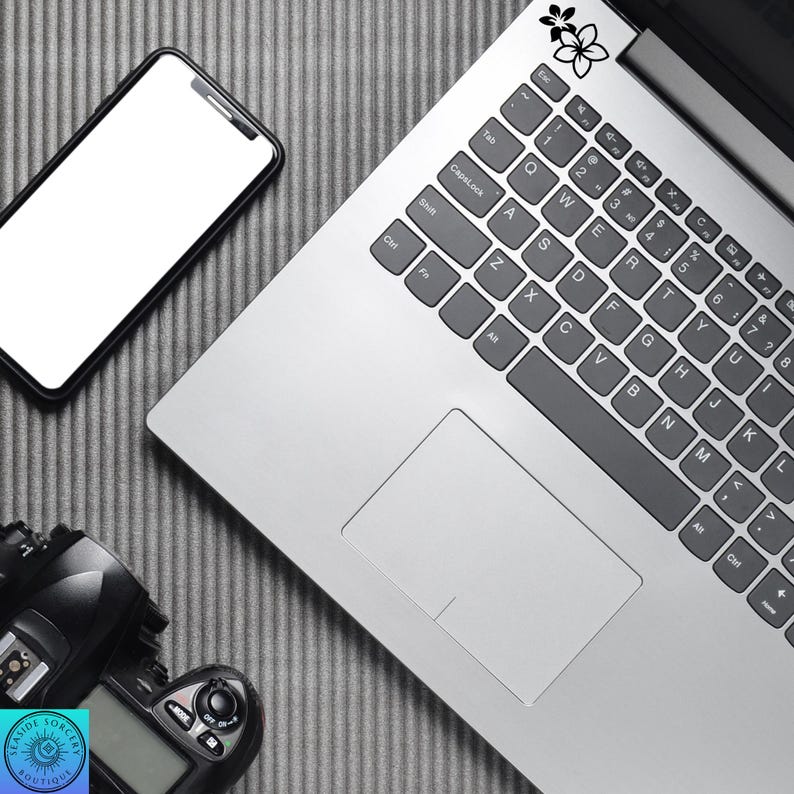 Pu&ograve; includere: Un laptop con un adesivo floreale in bianco e nero, uno smartphone e una fotocamera digitale sono disposti su una superficie grigia strutturata. La tastiera del laptop &egrave; visibile e la fotocamera ha un logo Seaside Sorcery Boutique.