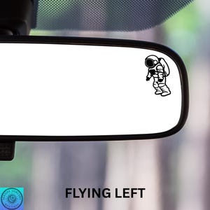 May include: Black and white astronaut sticker on a car rearview mirror. The sticker shows an astronaut in a spacesuit, with the text "FLYING LEFT" below the mirror.