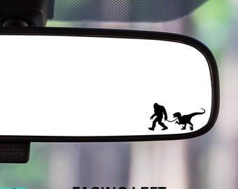May include: A black vinyl decal featuring a silhouette of Bigfoot walking a T-Rex on a leash, positioned on a car's rearview mirror. The text "FACING LEFT" is below the mirror.