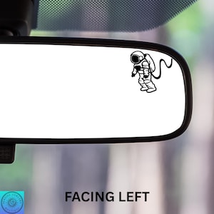 May include: Black and white astronaut decal on a car rearview mirror. The astronaut is facing left, with a detailed spacesuit design and a curved hose. The text "FACING LEFT" is below the mirror.