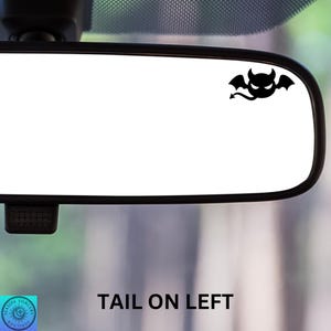 May include: A black silhouette of a devil with wings and a tail is on a white rearview mirror. The text "TAIL ON LEFT" is printed below the mirror.