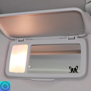 May include: A white car visor with a built-in mirror and a small light. A black and white French Bulldog decal is on the mirror. The visor is open, and the light is on.