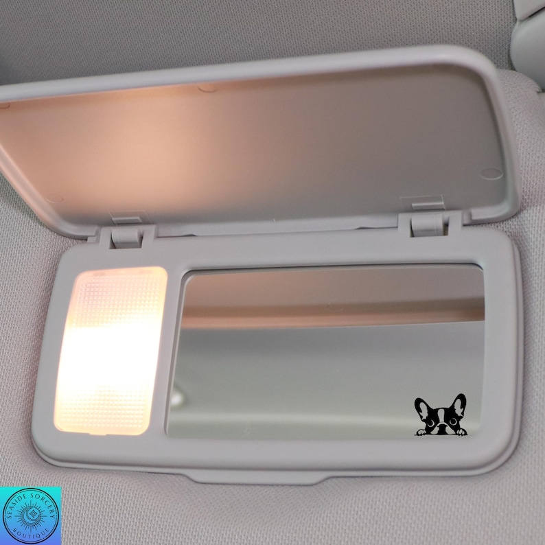 May include: A white car visor with a built-in mirror and a small light. A black and white French Bulldog decal is on the mirror. The visor is open, and the light is on.