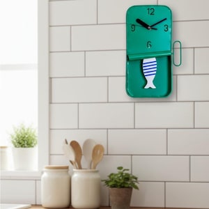 May include: A green metal wall clock with a fish design. The clock has black hands and numbers. The fish is white with blue stripes. The clock is mounted on a white tiled wall. The clock is approximately 25 cm tall.