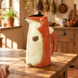 May include: A ceramic pitcher shaped like a fox with a glossy orange back and tail, a cream-coloured front, and small brown paws. The fox head forms the spout of the pitcher, featuring pointed ears and a dark nose, with a handle attached to the back.