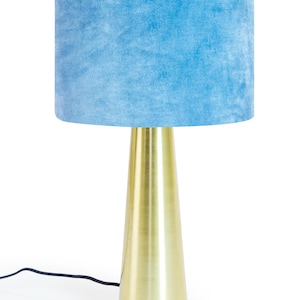 May include: A contemporary table lamp featuring a light blue velvet lampshade and a gold-coloured metal base. The base has a conical shape. A black power cord extends from the base. The lamp is set against a white backdrop.
