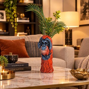 May include: A ceramic vase shaped like an orangutan head and torso, featuring a vibrant orange and deep blue glaze. The sculpture has a cylindrical base and holds a green leafy plant with small yellow berries, creating a striking focal point.