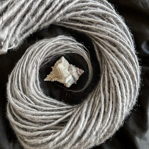 May include: A close-up of a bundle of silver grey synthetic dreadlocks. The dreadlocks are arranged in a circle with a seashell in the centre.