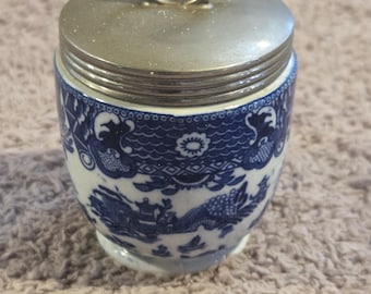 Op de afbeelding: Een keramische eierkoker met een blauw-wit willow-patroon. De houder heeft een metalen, zilverkleurig deksel met een cirkelvormige ringgreep aan de bovenkant, ontworpen om het deksel vast te zetten tijdens het koken van een ei.