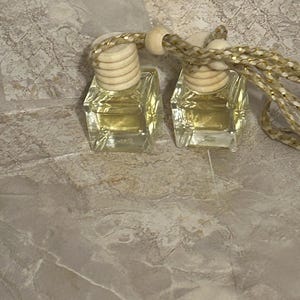 Könnte beinhalten: Zwei kleine, quadratische Glasflaschen, gefüllt mit einer gelben Flüssigkeit. Jede Flasche hat einen Holzverschluss und eine dekorative gold-beige Schnur zum Aufhängen. Wahrscheinlich Auto-Lufterfrischer oder Diffusoren.