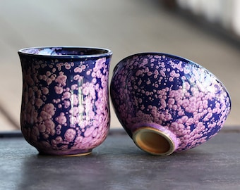 May include: Two ceramic teacups with a blue and purple speckled glaze. The cups are both round and have a wide opening.