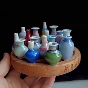 以下が含まれることがあります： 様々な色とデザインのミニチュアセラミックの花瓶のコレクションが、木製の台座に飾られています。花瓶は円形に配置され、台座は中央にあります。台座には「XINYU」という言葉が刻まれています。