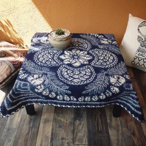 Könnte beinhalten: Eine blaue und weiße Batik-Tischdecke mit einem Blumenmuster. Die Tischdecke liegt auf einem quadratischen Holztisch mit schwarzen Beinen. Eine kleine Topfpflanze steht auf dem Tisch.