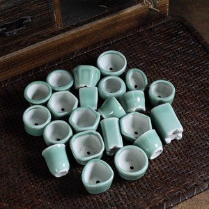 Può includere: Una collezione di piccoli vasi in ceramica verde chiaro di varie forme, tra cui rotondi, esagonali e rettangolari. Ogni vaso ha un interno bianco e un foro di drenaggio. I vasi sono disposti su una superficie tessuta.
