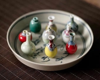 May include: A collection of small, colorful ceramic vases arranged on a white plate with a blue and white design. The vases are various shapes and sizes, with some featuring intricate patterns.