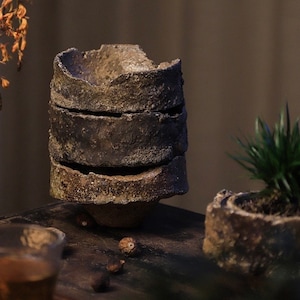 Puede incluir: Dos macetas de cerámica rústicas de color marrón con superficies rugosas e irregulares. La maceta más grande está apilada en tres secciones y la maceta más pequeña tiene una pequeña planta verde en su interior.