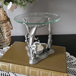 May include: A silver-toned metal oil burner featuring three sculpted dolphins rising from waves as a base, supporting a clear glass dish at the top. A small white tea light candle sits in the centre of the metal dolphin structure.