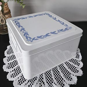 Pode incluir: Uma caixa de armazenamento retangular de metal branco com um design floral azul na tampa. A caixa está em uma toalha de renda branca. Um vaso de flores cor-de-rosa está ao fundo.