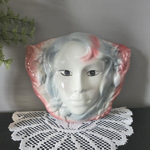 May include: A ceramic wall hanging shaped like a face. The face is white with black eyes, grey hair, and pink accents. The wall hanging is displayed on a black surface with a white lace doily.