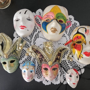 Pode incluir: Uma coleção de máscaras decorativas de cerâmica, com vários designs e cores. As máscaras incluem um estilo bobo da corte com acabamentos dourados, uma máscara com um design colorido e uma máscara com um rosto triste. As máscaras estão dispostas sobre uma toalha de renda branca.