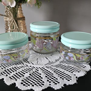 May include: Three clear glass storage jars with light mint green plastic lids. Each jar features a delicate floral pattern with pink and blue flowers, green leaves, and a thin gold band encircling the middle. The jars have a rounded shape.