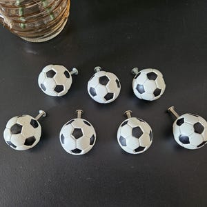 May include: A set of seven spherical cabinet knobs designed to resemble footballs, featuring the classic black and white pentagonal and hexagonal pattern. Each knob is attached to a short, silver-toned metal screw for mounting purposes.