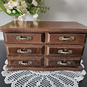 May include: A dark brown wooden jewellery chest featuring six small drawers arranged in two columns. Each drawer is equipped with an ornate, antique-style brass-coloured metal handle. The chest has a smooth, rectangular top surface and a base.