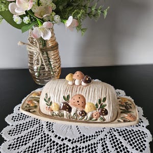 May include: A ceramic butter dish with a textured, wood-grain appearance, featuring a lid topped with three small sculpted mushrooms. The base and lid are decorated with painted mushroom clusters in shades of brown, yellow, and orange with green leaves.