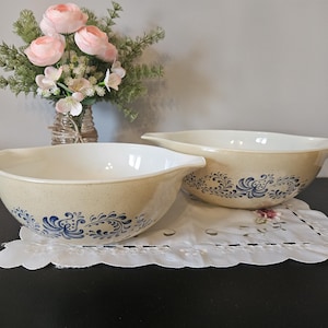 May include: Two beige mixing bowls with blue floral patterns. The bowls have handles and are on a white doily. A vase of pink flowers is in the background.