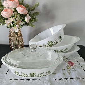 Könnte beinhalten: Ein Set weißer, ovaler Auflaufformen mit grünem Blumendesign. Eine Form hat einen Deckel aus klarem Glas mit einem Knauf. Die Formen sind auf einer weißen, bestickten Deckchen gestapelt. Im Hintergrund eine Vase mit rosa Blumen.