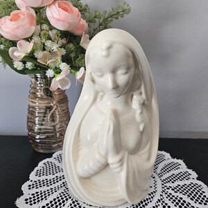 以下が含まれることがあります： 祈りのポーズをした女性のクリーム色のセラミック像。像は穏やかな表情をしています。ピンクの花が入った花瓶と白いレースのドイリーを背景にしています。