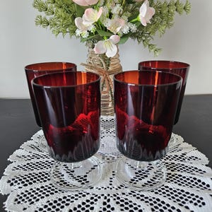 May include: Four ruby red drinking glasses with clear bases are arranged on a white lace doily. A vase of pink flowers and greenery sits in the background. The glasses have a vintage aesthetic.