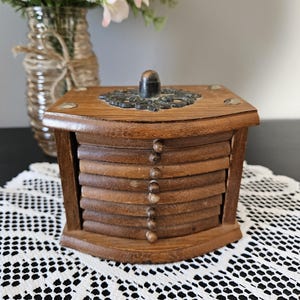 May include: A wooden coaster set with a decorative metal top and a stack of coasters. The coasters are made of wood and have a natural finish. The set is displayed on a white lace tablecloth.