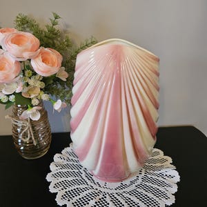 Könnte beinhalten: Eine Keramikvase in Form einer großen Muschel mit abwechselnden vertikalen Streifen in zartem Rosa und Creme. Die Oberfläche weist eine gerippte, kannelierte Textur auf, die die natürlichen Rillen einer Jakobsmuschel nachahmt, mit einer weiten Öffnung.